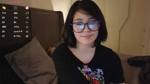 Media: Video of a young woman with short black hair, wearing glasses, a black T-shirt, and sitting on a bed with beige pillows. Background shows a phone screen displaying app details.