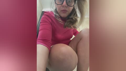 Media: A video of a fair-skinned woman with shoulder-length blonde hair, wearing black-rimmed glasses and a tight red sweater, sitting with her legs crossed, showing a large, round, pregnant belly.