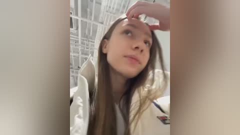 Media: Video of a young Caucasian woman with long brown hair, wearing a white jacket, touching her forehead in a reflective manner, in a modern, possibly indoor setting with white metal bars in the background.