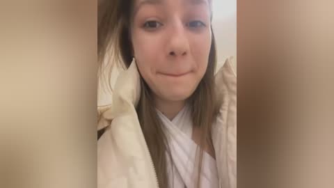 Media: Video of a young Caucasian woman with fair skin, brown hair, and a slight smile, wearing a white shirt and beige jacket, taken indoors with a soft, warm lighting.