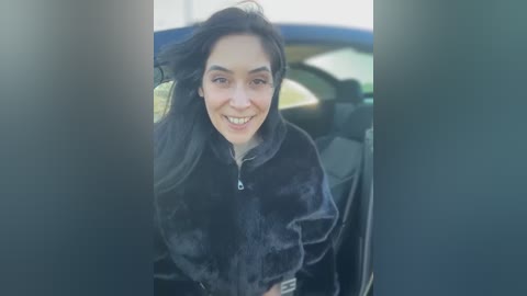 Media: Video of a smiling woman with long black hair and olive skin, wearing a black fur-lined jacket, standing inside a car with a blue car visible outside.