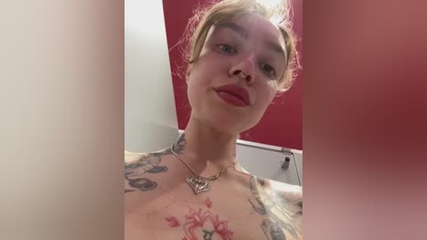 Media: Video of a pale-skinned woman with short blonde hair, wearing red lipstick, tattoos on chest and neck, and a silver necklace. Background features a red wall and white door.