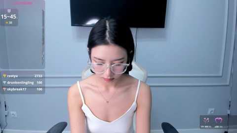 Media: Video of an Asian woman with pale skin and straight black hair, wearing glasses, a white camisole, sitting indoors.