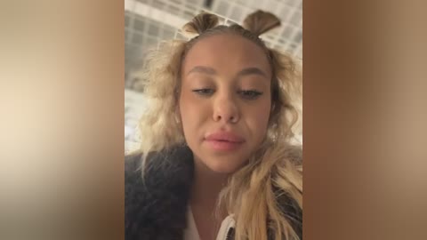 Media: Video of a young woman with light brown skin, curly blonde hair, and full lips, wearing a black fur-trimmed jacket. She gazes intently into the camera, set against a blurred, beige-tiled background.