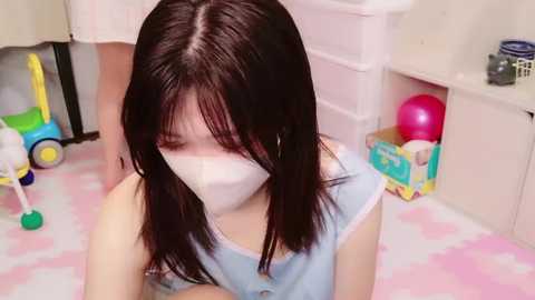 Media: A video of an Asian girl with straight black hair and light skin wearing a blue dress and a white mask, kneeling on a pink and white patterned rug in a bright, organized room with toys and furniture.