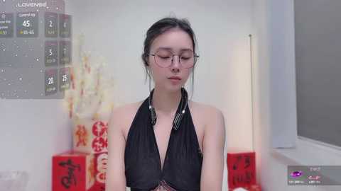 Media: Video of an Asian woman with fair skin and glasses, wearing a black halter top, standing in a minimalist room with a calendar, floral arrangement, and a window.