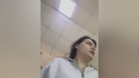 Media: A video captures a young woman with fair skin and long dark hair, wearing a white shirt, standing in a beige-tiled bathroom with a fluorescent light overhead.