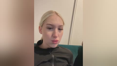 Media: Video of a young woman with fair skin and straight, blonde hair tied back, wearing a black jacket. She sits on a teal cushion in a simple, neutral-toned room. Her expression is neutral, and she appears to be looking down.