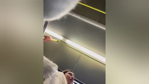 Media: Video of a woman in a white sweater, standing indoors near a bright yellow ceiling light. The image is slightly blurred, with a red object, possibly a balloon, partially visible in the background.