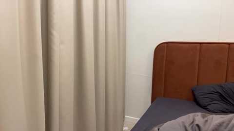 Media: Video of a minimalist bedroom corner with beige curtains covering a window, a brown leather headboard, and a dark gray bed with wrinkled sheets and a pillow.