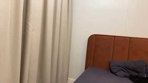 Media: A video of a minimalist bedroom with beige curtains, a brown padded headboard, and a dark gray bedsheet. The room is sparsely furnished with a single bed in the corner, giving a clean, modern aesthetic.