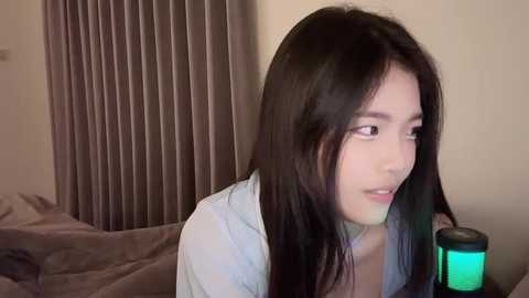 Media: Video of a young East Asian woman with long black hair, light skin, and delicate features, wearing a light blue shirt, sitting on a bed with brown sheets, next to a glowing green lamp, in a dimly lit room.