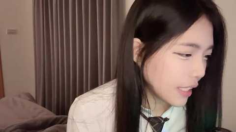 Media: Video of an Asian woman with long black hair, wearing a light-colored shirt, looking downward. Background features a bed with gray sheets and vertical beige curtains. Soft lighting creates a warm ambiance.