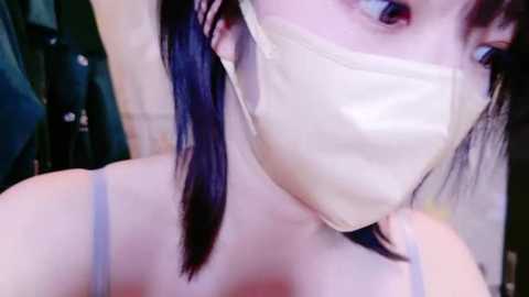 Media: A close-up video of an Asian woman with long, straight, dark hair, wearing a beige face mask, revealing part of her eyes and collarbone. The background is blurred, showing a green towel and a dark wall.