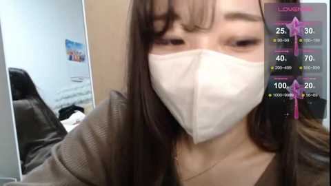 Media: A close-up video of a woman with long brown hair, wearing a white surgical mask and brown sweater, taken in an office setting with a digital overlay showing health statistics.