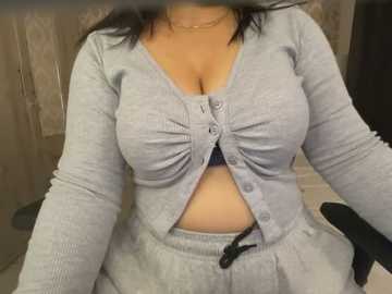 Media: Video of a woman in a tight, light gray, ribbed, long-sleeved top with a button-down front, showcasing ample cleavage. She wears matching gray sweatpants and sits on a black office chair. Background shows a beige wall and wooden door.