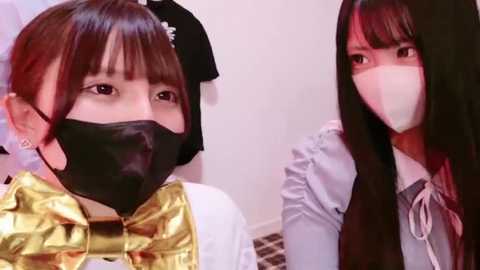 Media: Video of two Asian women in masks, one with straight black hair, the other with straight brown hair. Both wear gold metallic tops. Background shows a simple, white-walled room with clothes hanging.