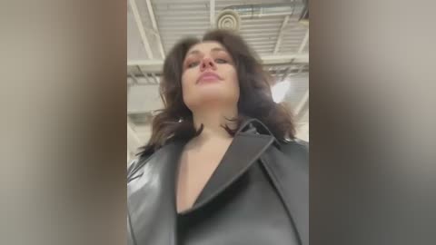 Media: Video of a young woman with fair skin, wavy dark hair, and a confident expression, wearing a black leather jacket, standing in an industrial, high-ceilinged room with exposed pipes and ductwork.