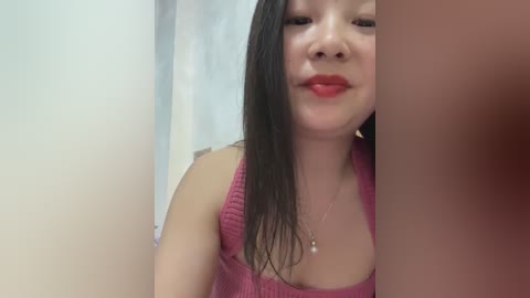 Media: Video of an East Asian woman with long black hair, wearing a pink tank top, smiling, in a bathroom with a white towel in the background.