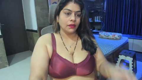 Media: Video of a South Asian woman with long black hair, wearing a maroon bra, sitting in a dimly-lit room with a kitchen in the background.