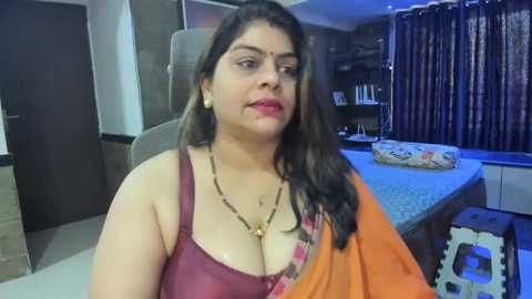 Media: Video of a South Asian woman with long dark hair, fair skin, and medium build, wearing a maroon bra and orange sari, seated in a modern, dimly lit room with a kitchen counter, dark curtains, and a closed door.