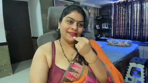 Media: Video of an Indian woman with medium-dark skin, wearing a pink sari with orange borders, sitting on a gray sofa, smiling, in a modern, well-lit living room with a blue-patterned bedspread and dark curtains.