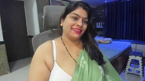 Media: Video of a South Asian woman with medium brown skin, long black hair, and a green saree, sitting on a grey chair in a modern, dimly lit bedroom with dark curtains and a bed with a patterned duvet.