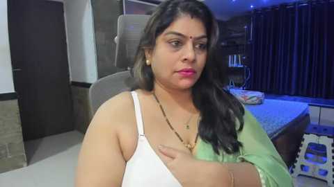 Media: Video of a middle-aged woman with long black hair, wearing a white sleeveless top and green sari, sitting on a beige chair in a dimly lit bedroom with dark curtains and a bed.