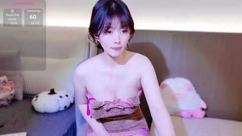 Media: Video of a young Asian woman with dark hair, wearing a pink and brown striped tube top, sitting on a bed with plush toys in a modern, softly lit room.