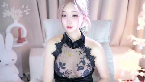 Media: Video of an Asian woman with fair skin and pink hair, wearing a sheer black lace dress, seated on a white couch. Background features white decor, including a bunny statue and fairy lights.