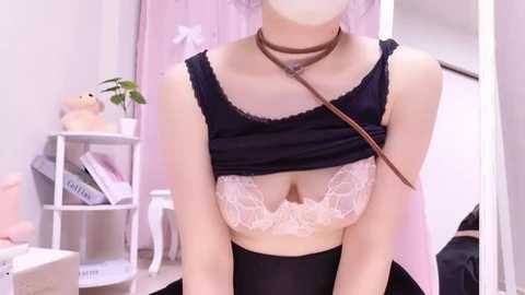 Media: Video of a slender, fair-skinned woman with light brown hair, wearing a black sleeveless top lifted to reveal a lace bra with pink floral patterns, and a brown choker. Background features a white shelf with books, teddy bear, and a pink curtain.