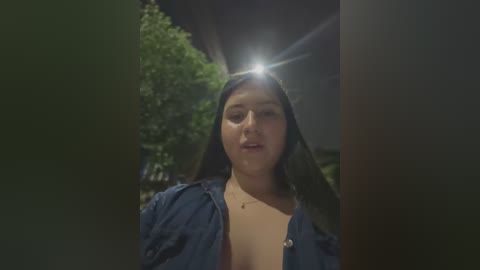 Media: Video of a young woman with long black hair and fair skin, wearing a blue denim jacket and a low-cut top, standing outdoors at night.