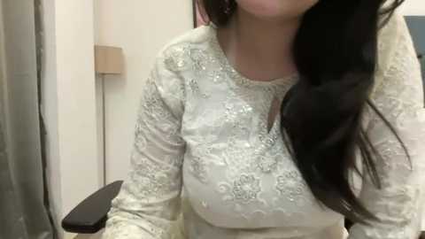 Media: Video of a woman with long, dark hair wearing an elegant, white, long-sleeved, lace-trimmed dress with intricate floral embroidery. She is sitting in a modern, light-colored room with a beige wall and a black chair.