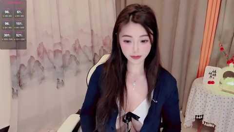 Media: Video of a young East Asian woman with long dark hair, wearing a blue blazer over a white lace bra, sitting in a chair in a softly lit room with floral curtains.