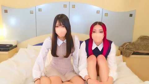 Media: Video of two young Asian women with long hair, sitting on a bed in a modern hotel room. They wear white shirts and striped overalls, with one in red hair, and the other in black hair.