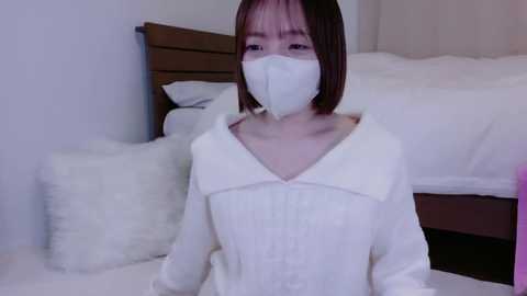 Media: Video of an East Asian woman with short dark hair, wearing a white face mask and long-sleeved white shirt, kneeling on a white bed in a minimalist bedroom with a wooden headboard and fluffy pillows.