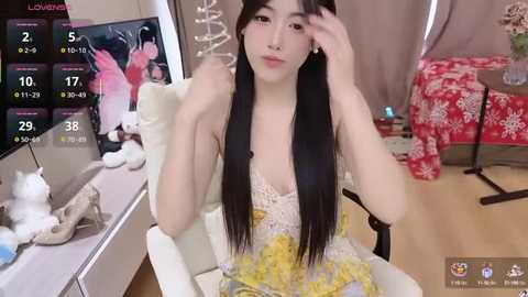 Media: Video of an Asian woman with long black hair, light skin, and a slender figure, wearing a yellow floral dress, sitting on a white chair, with a TV displaying a live stream, in a cozy, decorated room.