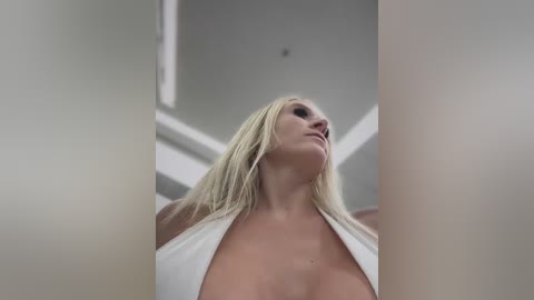 Media: A video of a blonde woman in a white halter top, emphasizing her large breasts, standing in a modern, white-lit room. The image is cropped, focusing on her upper body and face, with a slightly blurry background.