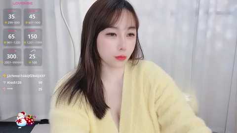 Media: Video of a young East Asian woman with fair skin, long dark hair, and red lipstick, wearing a yellow bathrobe, sitting in a room with white curtains.