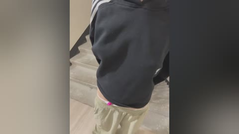 Media: Video of a person in a black hoodie and light green sweatpants, standing on a tiled floor, with a pink object visible at the waistband.