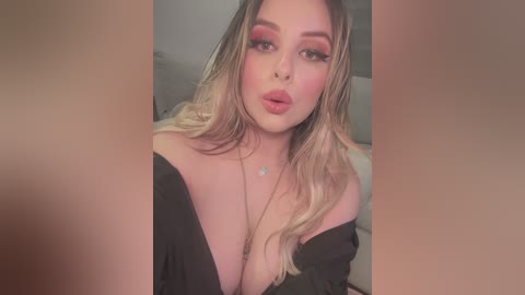 Media: Video of a fair-skinned woman with long, wavy blonde hair, wearing bold makeup, including red lipstick and dramatic eye makeup. She has a plunging black off-shoulder top, revealing ample cleavage, and is captured indoors, possibly in a bedroom setting.