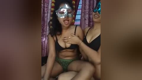 Media: A video of two women in a dimly lit room, wearing black bras and green lace panties, with silver masquerade masks, one covering her face, the other smiling.