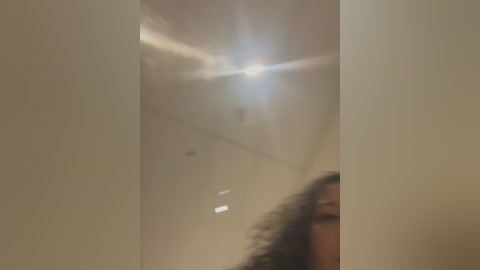 Media: A blurred video shows a person with curly hair, partially visible, standing in a dimly lit room with a bright light source creating lens flares. The background features off-white walls and a few ceiling fixtures.