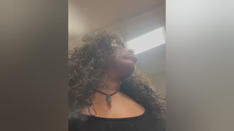 Media: A video of a young woman with curly hair, wearing a black top, standing in a dimly lit corridor. She has a necklace with a green pendant.