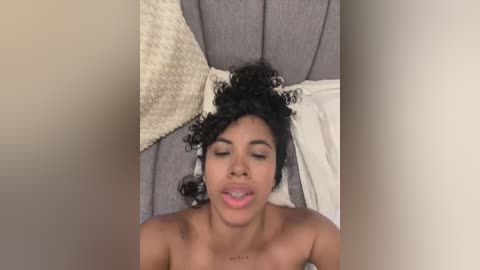 Media: Video of a topless Black woman with curly hair, lying on a gray couch, with a beige pillow and white blanket in the background.