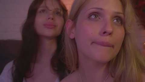 Media: Video of two young women with fair skin and light hair, one with bangs, the other with straight hair, looking at each other, indoor setting with soft lighting.