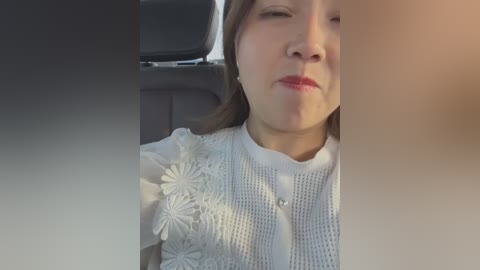 Media: Video of an Asian woman with fair skin, light brown hair, and a white, textured blouse with floral embroidery, sitting in a car with a blurred background.