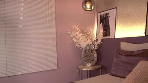 Media: Video of a cozy bedroom corner with lavender walls, white blinds, a beige bed, a gold vase with dried branches, and a framed black-and-white photo on the wall.