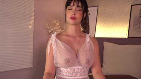 Media: A video of a Caucasian woman with dark hair, wearing a sheer, lace bralette, standing in a dimly-lit room with beige walls and blinds.