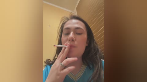 Media: Video of a young woman with long brown hair, wearing a blue top, smoking a cigarette, standing in a room with yellow walls and wooden blinds.